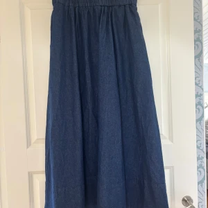 Blå långkjol i denim från Neo Noir - Snygg långkjol i mörkblå denim från Neo Noir. Klänningen har en rak och luftig passform med resår i midjan för extra komfort. Perfekt för dig som gillar en enkel men trendig look. Materialet är jeans och färgen är klassiskt mörkblå.