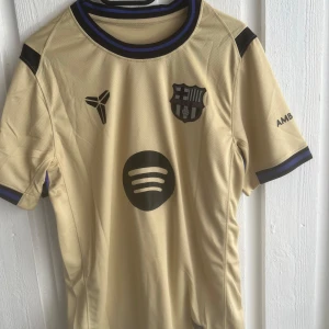 FC Barcelona Lamine Yamal fotbollströja - Snygg FC Barcelona fotbollströja i beige med svarta detaljer och lila/blå ränder vid krage och ärmslut. Tryck med Lamine Yamal och nummer 10 på ryggen, samt flera officiella loggor som Spotify och UNHCR. Tillverkad i lätt och ventilerande material.
