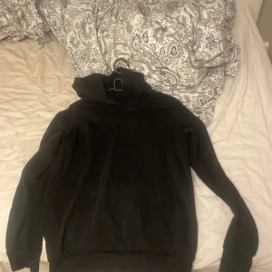 Svart hoodie från Peak Performance - Svart hoodie från Peak Performance med broderad logga på bröstet. Klassisk modell med huva och känguruficka framtill. Mjuk bomullskänsla och långärmad, perfekt för chill dagar eller när du vill ha en clean look. Storlek 160 så passar xs/xxs