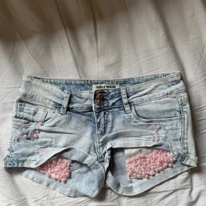 Ljusblå jeansshorts med pärlor Tally Weijl - Supersnygga ljusblå jeansshorts från Tally Weijl med slitna detaljer och rosa pärlor på framsidan. Shortsen har låg midja, klassiska fickor och coola rivna partier. Perfekta för sommaren och ger en unik touch till din outfit!