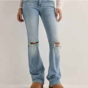 Ljusblå bootcut jeans med slitningar - Snygga ljusblå jeans med bootcut passform och slitna hål över knäna. låg midja och klassisk femficksmodell med dubbla knappar i midjan. Perfekta för en avslappnad och trendig look.