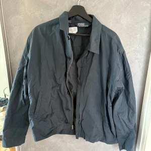 Mörkblå vindjacka från Polo Ralph Lauren - Snygg mörkblå vindjacka från Polo Ralph Lauren i klassisk modell. Jackan har dragkedja och knappar framtill, två fickor och en stilren krage. Perfekt för dig som gillar enkel och clean stil. Materialet är lätt och känns skönt att bära under vår och höst.