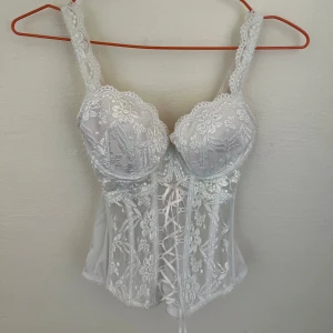 Vit spetskorsett med snörning 34B - Superfin vit korsett med blommig spets och snygg snörning framtill. Justerbara axelband och hakknäppning bak. Perfekt för dig som vill ha en romantisk och feminin vibe. Materialet är polyester och elastan för skön passform.