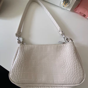 Beige axelväska från Gina Tricot - Snygg beige axelväska från Gina Tricot med croco-mönstrad yta och silvriga metalldetaljer. Väskan har en avtagbar axelrem och stängs med dragkedja. Perfekt storlek för det viktigaste och ger en trendig touch till din outfit.