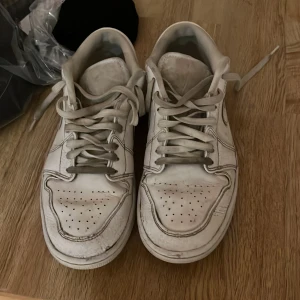 Vita Nike Air Jordan 1 sneakers - Klassiska vita Nike Air Jordan 1 sneakers med perforerad tå och ikonisk Air Jordan-logga på hälen. Skorna har snörning och är tillverkade i skinn, perfekt för dig som gillar streetstyle och vill ha en tidlös look.