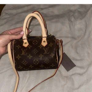 Mini handväska  - Superfin liten handväska med klassiskt monogrammönster i brunt och beige. Väskan har både handtag och axelrem, guldfärgad dragkedja och detaljer i ljust äkta läder. Perfekt för dig som vill ha en ikonisk accessoar med lyxig känsla