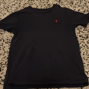 Mörkblå t-shirt från Polo Ralph Lauren - Svart t-shirt från Polo Ralph Lauren med klassisk röd broderad logga på bröstet. Enkel och stilren design med rund hals och korta ärmar. Perfekt basplagg för dig som gillar en clean look och vill ha något som funkar till allt.