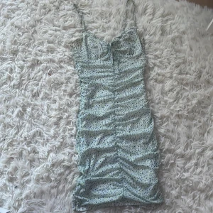 Blommig kortklänning från H&M Divided - Supersöt kortklänning från H&M Divided i blommigt mönster med tunna axelband och rynkade detaljer. Klänningen är ljus med små blå och gröna blommor, har figurnära passform och är perfekt för sommarens varma dagar.