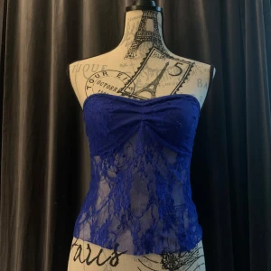 Blå spets bandeau tubtopp med knyt - Säljer en blå bandeau tubtopp i transparent spets med blommigt mönster. Toppen är axelbandslös och har snygg knytning i ryggen för perfekt passform. Perfekt för dig som vill sticka ut med en trendig och feminin look. Knappt använd