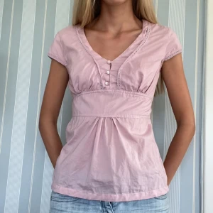 Ljusrosa blus med knyt och knappar - Supersöt ljusrosa blus med v-ringning, knappar framtill och knytband i ryggen. Korta ärmar och lätt rynk under bysten ger en feminin siluett. Perfekt till jeans eller kjol! Superfint skick💕