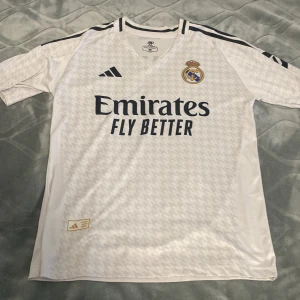 Real Madrid Mbappé 9 fotbollströja M - Vit Real Madrid fotbollströja med Mbappé och nummer 9 på ryggen. Tröjan har korta ärmar, svarta detaljer och klubbmärke på bröstet. Tillverkad i lätt och ventilerande material, perfekt för match eller träning. Snyggt subtilt mönster över hela tröjan.
