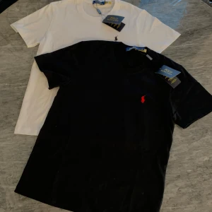 2-pack t-shirts Polo Ralph Lauren - Två klassiska t-shirts från Polo Ralph Lauren, en svart med röd broderad logga och en vit med svart broderad logga. Båda är i mjuk bomull, har rund hals och kort ärm. Perfekt basic-plagg med stilren design och ikonisk logga på bröstet. PASSAR ÄVEN XS