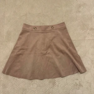 Kjol - Kjol från H&M i mjuk mockaimitation. Kjolen har A-linjeformad passform, dekorativa metallringar framtill och dragkedja bak. Storlek 42!