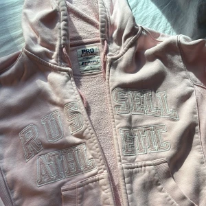 Ljusrosa hoodie från Russell Athletic - Säljer en ljusrosa hoodie från Russell Athletic med broderad text på bröstet. Tröjan har huva, dragkedja framtill och två fickor. Mjuk insida och klassisk collegekänsla, OBS dragkedjan funkar inte!
