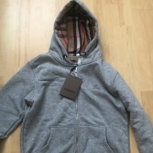 Grå burberry hoodie - Grå hoodiejacka från Burberry med dragkedja, klassiskt rutigt foder i huvan och broderad logga på bröstet. Jackan har snörning i huvan och två fickor framtill. Perfekt för dig som gillar stilren design med en touch av lyx. Den är helt ny och aldrig använt taggen är fortfarande kvar