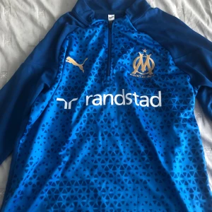 Blå långärmad Marseille fotbollströja Puma - Snygg långärmad fotbollströja från Olympique de Marseille i blått med geometriskt mönster framtill. Guldiga detaljer med klubbmärke och Puma-logga, samt vit Randstad-sponsor. Tröjan har halv dragkedja och är tillverkad i funktionsmaterial. Knappt använd och är i väldigt bra skick. Priset kan diskuteras!