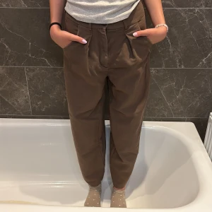 Bruna croppade kostym byxor med veck - Snygga bruna byxor med croppad längd och hög midja. Byxorna har veck framtill, sidofickor och bakfickor. De är i ett mjukt bomullstyg och har en relaxed, loose passform som ger en chill vibe. Men har ett litet hål i vänstra ficka. 