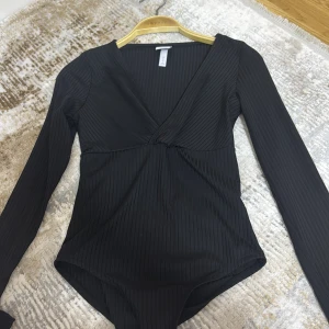 Svart omlott body från H&M - Svart ribbad body från H&M med omlott-skärning och djup v-ringning. Långärmad modell i stretchigt material. Storlek S