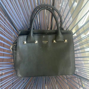 Svart handväska från Guess i skinn - Stilren svart handväska från Guess med två rejäla handtag och guldfärgade metalldetaljer. Väskan har flera fack, varav ett med dragkedja, och ljus insida med Guess-logga. Klassisk fyrkantig form i skinnimitation, perfekt för att bära allt du behöver. Äkta även i skinn. Finns små defekter se bilderna och även lite sminkfläckar inne  men som syns knappt❤️