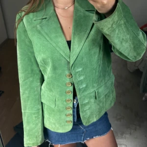 Äkta mocka jacka  - Äkta mocka jacka med klassisk krage och flera dekorativa knappar framtill. Jackan har två fickor med lock och en figurnära passform som ger en stilren look. Perfekt för dig som vill sticka ut med färg.