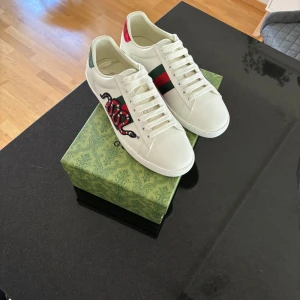 Gucci Ace  - Gucci Ace sneakers i vitt skinn med klassiska gröna och röda ränder på sidan. Ena skon har en broderad orm i rött, svart och vitt. Båda har metallicröd detalj på hälen. Snygg och lyxig modell med rund tå och snörning.