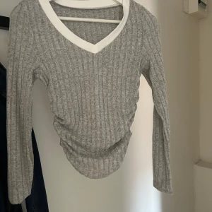 Grå ribbad långärmad tröja - Snygg grå ribbad tröja med lång ärm och vit v-ringad krage. Tröjan har en figurnära passform och är väldigt mjuk i materialet.