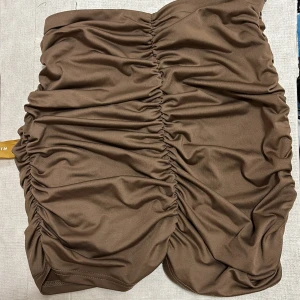 Brun kortkjol med rynkade detaljer - Säljer en brun kortkjol med snygga rynkade detaljer längs hela kjolen. Kjolen har en figurnära passform och är tillverkad i ett mjukt, stretchigt material som sitter bekvämt. Perfekt för dig som gillar trendiga och stilrena plagg.