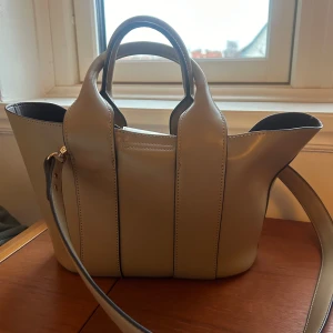 Beige axelväska i skinn med handtag - Snygg beige axelväska i slätt skinn med två rejäla handtag och avtagbar axelrem. Väskan har en stilren design med svarta kanter och ljus insida. Perfekt storlek för vardagens essentials och ger en clean look till din outfit.
