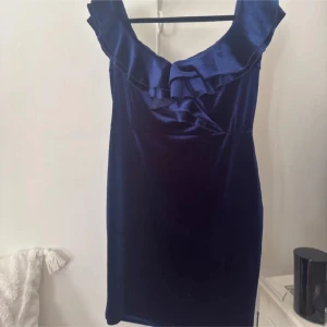 Mörkblå sammetsklänning med volang - Snygg mörkblå sammetsklänning i stretchig polyester med offshoulder och volangdetalj längs urringningen. Kort modell som sitter tight och ger en elegant look. Perfekt för dig som vill sticka ut med en klassisk men trendig stil. Storlek L men upplever den som en M.