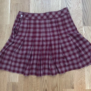 Rutig vinröd plisserad kortkjol - Supersnygg vinröd plisserad kortkjol med klassiskt rutigt mönster i vitt. Kjolen har en markerad linning och knäppning i sidan. Perfekt för en trendig och preppy look. Hör av dig om du vill ha fler bilder eller har några frågor! 