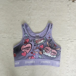 Lila sporttopp från Stronger med tryck - Supercool lila sporttopp från Stronger med färgglatt pop art-tryck, hjärtan och text som 'GRL PWR'. Bred resår med logga, brottarrygg och bekväm passform. Perfekt för träning och att sticka ut på gymmet.