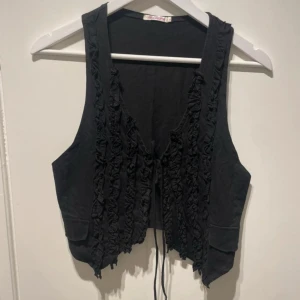 Svart väst/topp - Svart detaljerad väst/topp med ruffles och snören att knyta. Strl M.