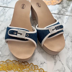 Scholl sandaler med denim och klack - Snygga sandaler från Scholl med blått denimtyg och vita detaljer. Skorna har en silverfärgad spänndetalj framtill, öppen tå och låg klack. Sulan är ljus och sandalerna har nitar på sidorna för extra edge. Perfekta för en chill sommarlook.