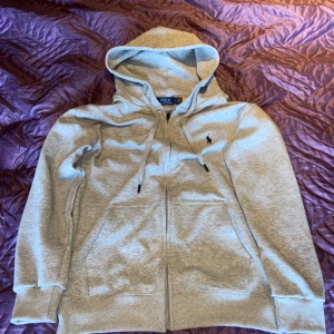 Polo Ralph Lauren - Snygg ljusgrå hoodie från Polo Ralph Lauren med dragkedja, huva och snörning. Klassisk logga broderad på bröstet och stora fickor framtill.