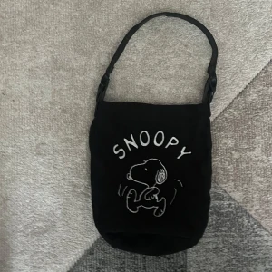 Svart Snoopy axelväska i tyg - Svart axelväska med broderad Snoopy-figur och text på framsidan. Väskan har en enkel, rundad form och ett brett axelband. Tillverkad i mjukt tygmaterial, perfekt för att bära dina vardagsgrejer och ge din outfit en lekfull touch.