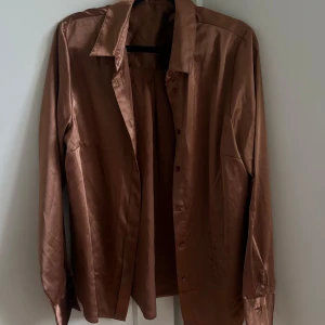 Brun satinliknande skjorta  - Säljer en rostbrun-ish skjorta från BikBok i satinliknande material. Skjortan har klassisk krage, långa ärmar och knäppning framtill. Perfekt för dig som gillar retrovibbar och vill sticka ut med en glansig finish. Snygg att styla både instoppad och lös. True to size. 