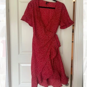 Röd omlottklänning med vita prickar - Superfin röd omlottklänning från Vero Moda med vita prickar, volangkant nertill och knyt i midjan. Kort ärm och v-ringad. Lätt och luftig modell. Går till knäna på mig (162 cm). Använd ett fåtal gånger.