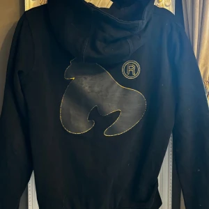 Svart Money hoodie med guldtryck - Svart hoodie från Money med stort guldigt logotryck på bröstet och en unik guldig detalj på ryggen. Tröjan har huva med snörning och långa ärmar. Perfekt för dig som gillar streetwear och vill sticka ut med snygga detaljer.