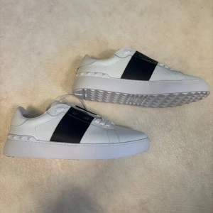Valentino Garavani vita sneakers - Snygga skor inspirerade av Valentino Garavani Open sneakers i vitt skinn med bred svart läderdetalj över vristen. Klassisk rund tå, vita snören och ikoniska nitar baktill. Perfekt för dig som vill ha en clean och lyxig look med streetkänsla. Skorna är Reeppss