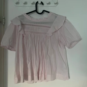 Ljusrosa blus med volang från Gina Tricot - Supersöt ljusrosa blus från Gina Tricot med volangdetaljer vid axlarna och rynkade partier. Blusen har korta ärmar och fina broderade band över bröstet. Perfekt för dig som gillar romantisk och luftig stil.