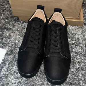 Svarta sneakers från Christian Louboutin - Stilrena svarta sneakers från Christian Louboutin med klassisk röd sula och diskreta detaljer. Skorna har snörning, rund tå och är tillverkade i en mix av mocka och skinn. Perfekta för dig som gillar lyxiga och minimalistiska vibes.