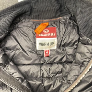 Svart dunväst från Parajumpers M - Snygg svart dunväst från Parajumpers med quiltad framsida, dragkedja och cool gul detalj vid halsen. Västen har flera fickor med dragkedja och klassisk Parajumpers-logga på ryggen. Perfekt lager-på-lager-plagg för kyliga dagar.