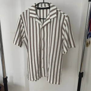 Randig kortärmad skjorta i bomull - Snygg skjorta med raka, vertikala ränder i beige och vitt. Skjortan har korta ärmar, klassisk krage och knäppning framtill. Perfekt för en chill och stilren look under varmare dagar. Materialet känns mjukt och luftigt.
