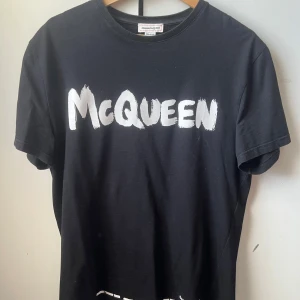 Alexander McQueen T-shirt - Svart t-shirt från Alexander McQueen med vit, stor McQueen-logga framtill. Klassisk rund halsringning och korta ärmar. Tillverkad i mjuk bomull och har en clean, stilren look som passar perfekt till jeans eller shorts. Äkta T-shirt