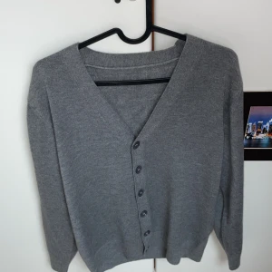 Grå tröja/overshirt - En stilren grå kofta med v-ringning och knäppning framtill. Koftan har långa ärmar och är gjord i ett mjukt, stickat material som passar perfekt för lager på lager. Det är en manlig tröja men den passar verkligen både könen