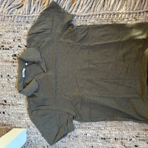 Mörk grön pikétröja från Bläck - Säljer en stilren mörk grön pikétröja från Bläck. Tröjan har klassisk krage, korta ärmar och är gjord i mjuk bomull. Perfekt för en clean och avslappnad look. Passar dig som gillar enkel och tidlös stil.