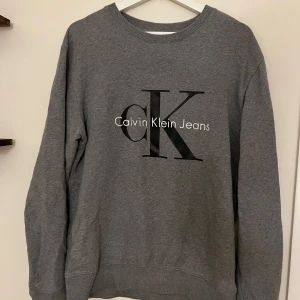 Grå sweatshirt från Calvin Klein Jeans - Snygg grå sweatshirt från Calvin Klein Jeans med stor svart CK-logga framtill. Klassisk rund halsringning och långa ärmar. Perfekt för en chill och stilren look. 