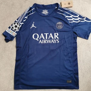 PSG x Jordan blå fotbollströja #7 - Snygg blå PSG fotbollströja med vita detaljer, Jordan-logga och Qatar Airways-tryck på bröstet. Unikt vingmönster på axlarna och Champions League-märke på ärmen. Nummer 7 och Kvaratskhelia på ryggen. Perfekt för dig som älskar fotboll och streetstyle. Den är äkta från Frankrike 