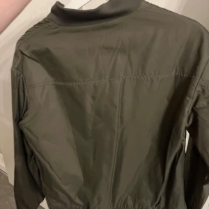 Olivgrön bomberjacka från NA-KD - Snygg olivgrön bomberjacka från NA-KD med klassisk ribbad krage, muddar och dragkedja framtill. Jackan har två fickor och är tillverkad i ett slätt syntetmaterial som ger en clean look. Perfekt för dig som vill ha en stilren och enkel jacka.