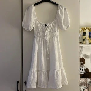 Vit kortklänning från Gina Tricot - Supersöt vit kortklänning från Gina Tricot med puffärmar, knytning framtill och volangkant nertill. Klänningen har en lätt skrynklig struktur och är perfekt för sommarens alla tillfällen. Sval och luftig modell med feminin passform💞💐